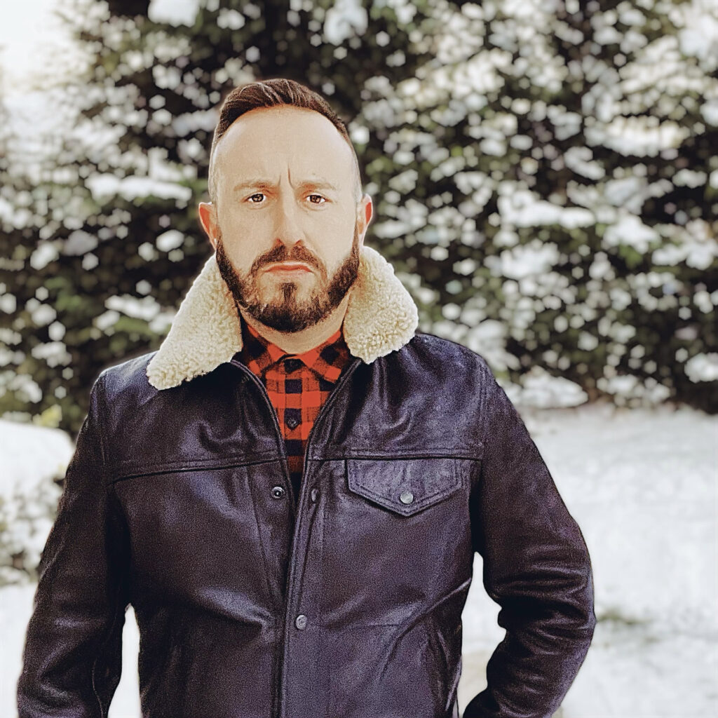 Man wearing a leather jacket, plaid shirt, and jeans standing in a snowy landscape with pine trees in the background.
